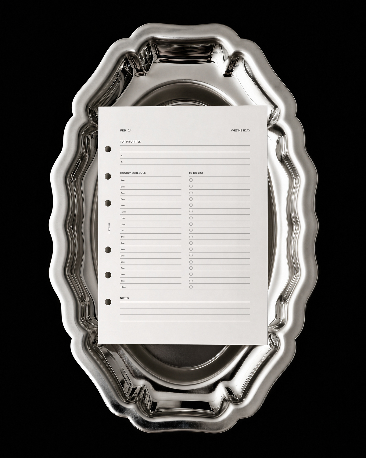 A white Notaire 2027 dated planner insert with black printed text and circular holes on the left side, placed on top of an ornate silver metal serving platter against a solid black background.