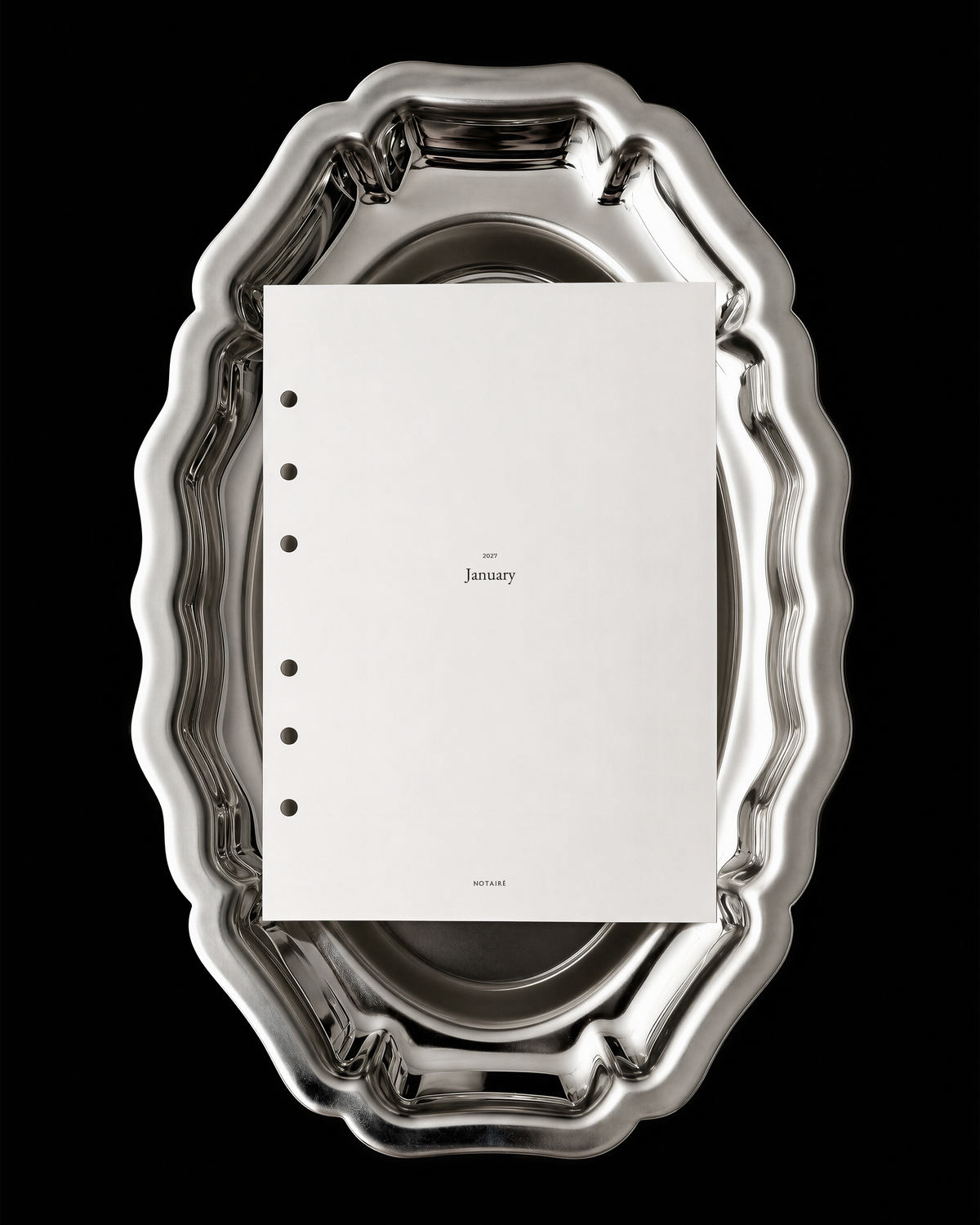 A white Notairé planner divider with black printed text and circular holes on the left side, placed on top of an ornate silver metal serving platter against a solid black background.