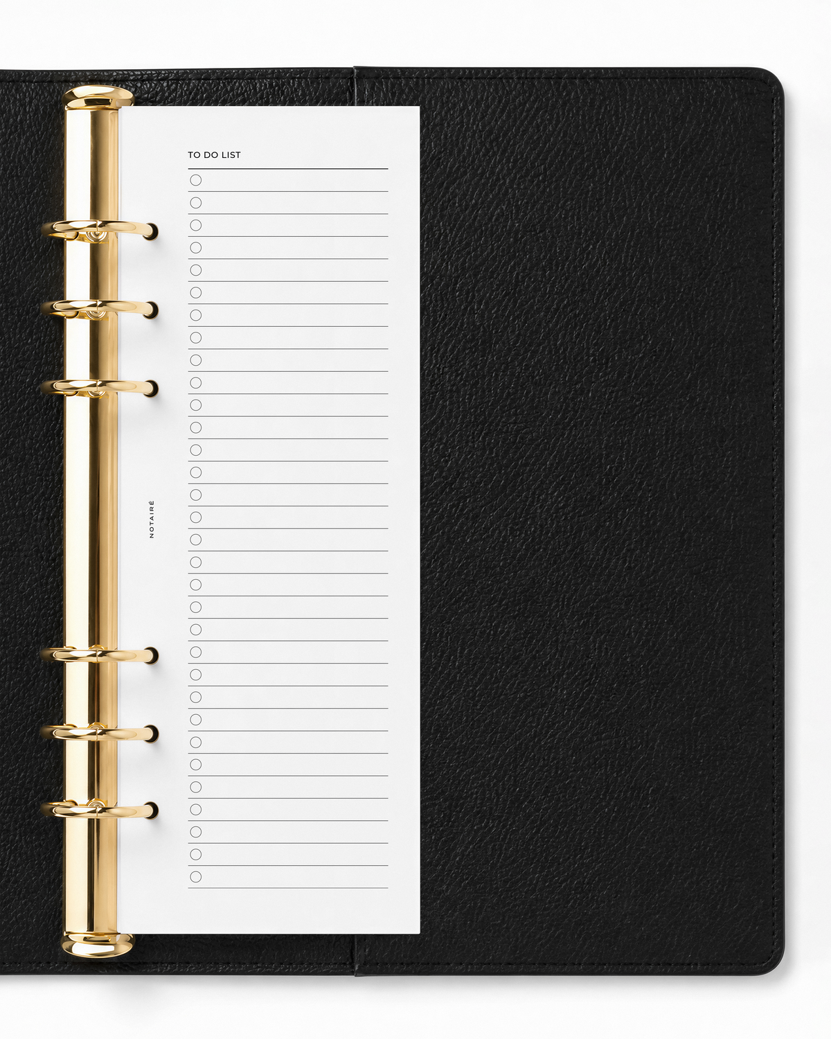 A black textured ring binder planner shown open, featuring a white paper half-page A5 Notairé insert with a shopping list template and gold-tone metal rings.
