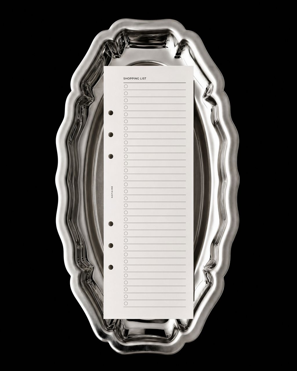 A white shopping list Notairé half-page insert with lined checkboxes sits centered inside an ornate, reflective silver metal tray against a solid black background.