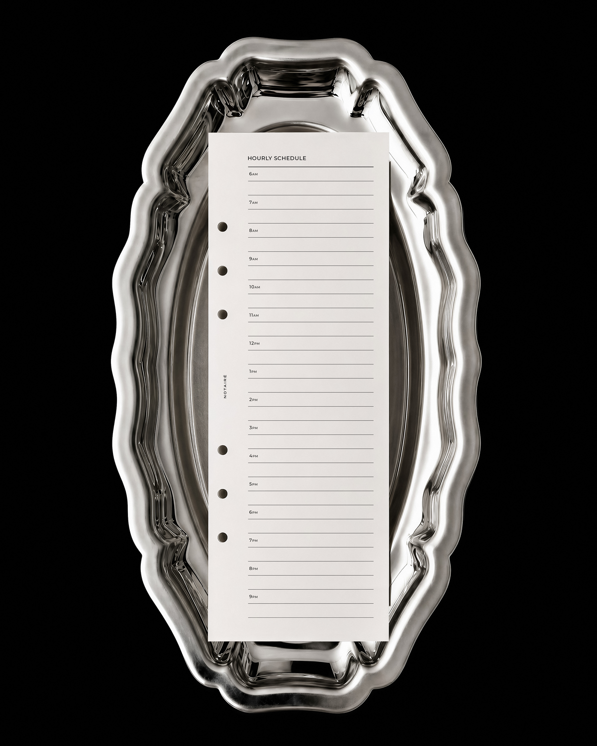 A white shopping list half-page planner insert from Notairé with lined checkboxes sits centered inside an ornate, reflective silver metal tray against a solid black background.