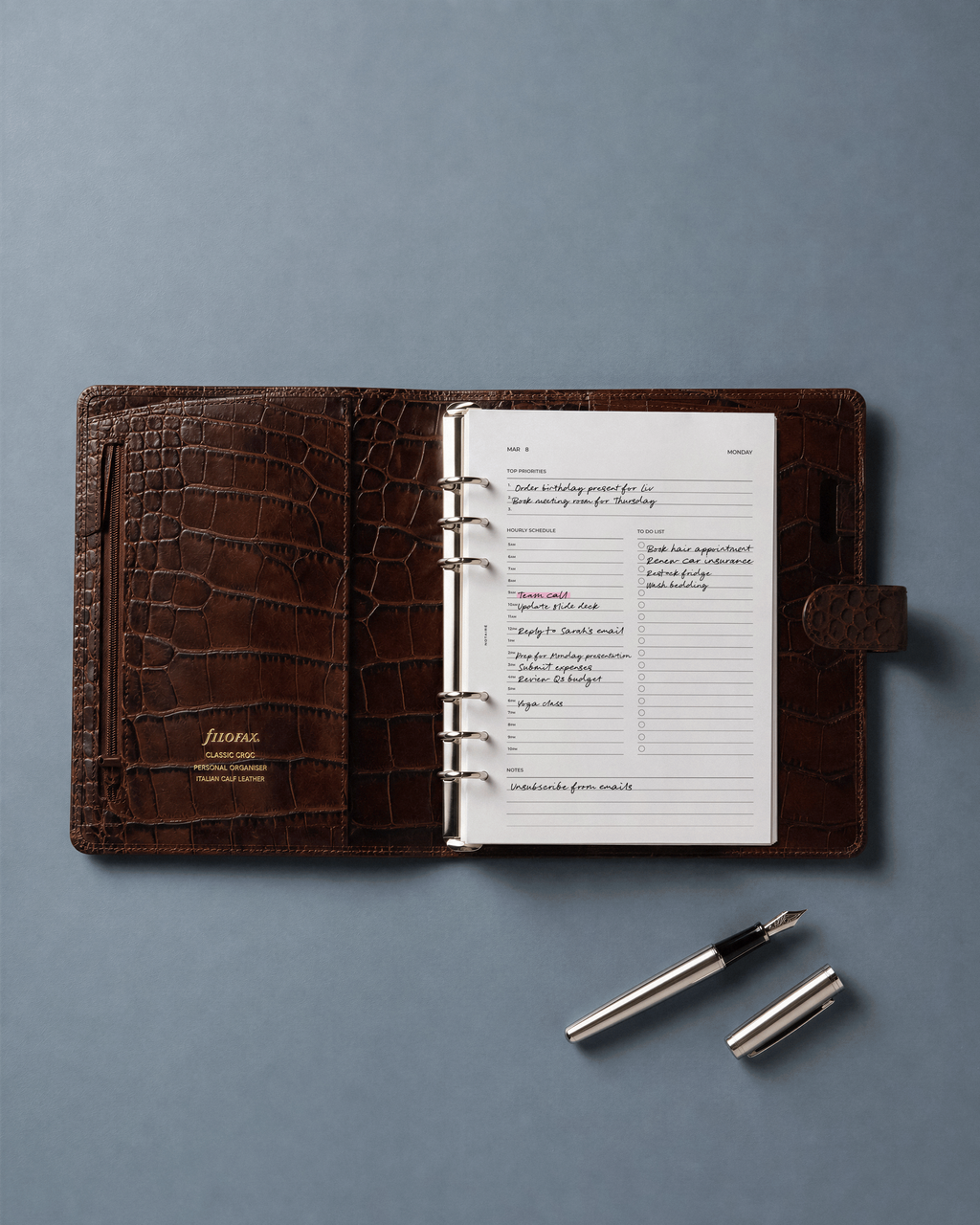 A brown crocodile-embossed ring-bound Filofax open on a muted blue-gray background, with a silver pen placed below it. With White Notaire dated 2027 daily planner inserts.