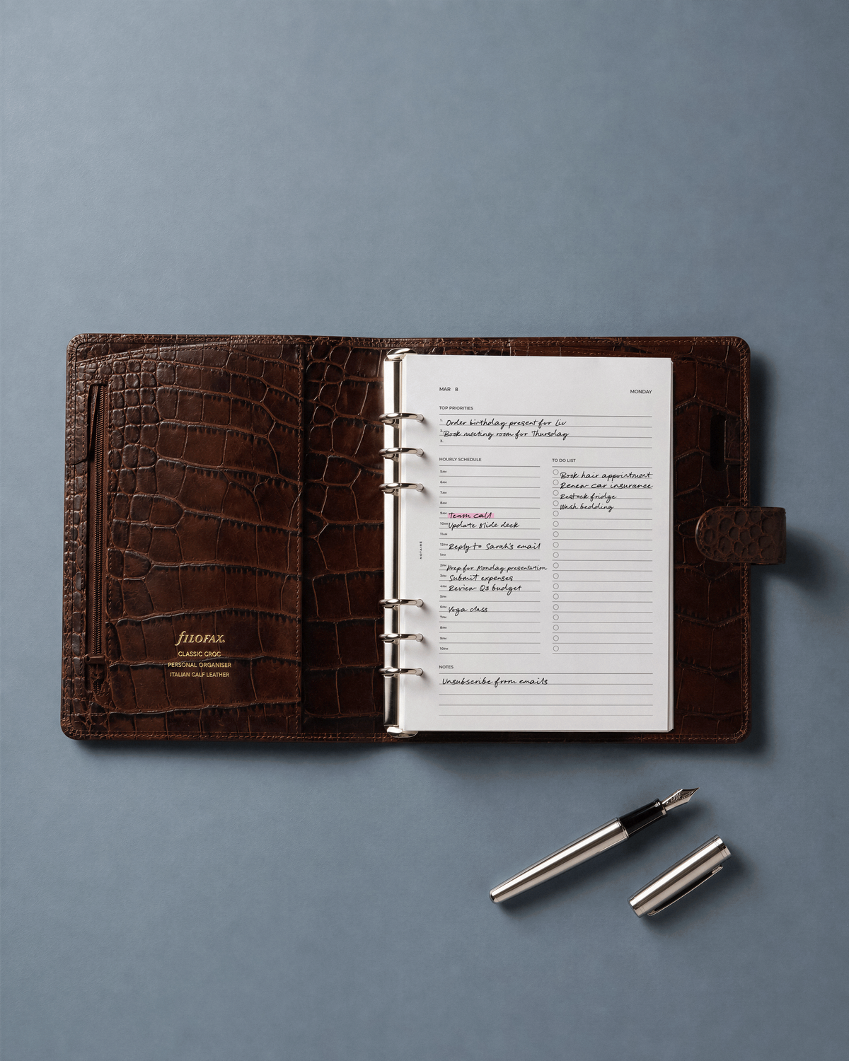 A brown crocodile-embossed ring-bound Filofax open on a muted blue-gray background, with a silver pen placed below it. With White Notaire dated 2027 daily planner inserts.