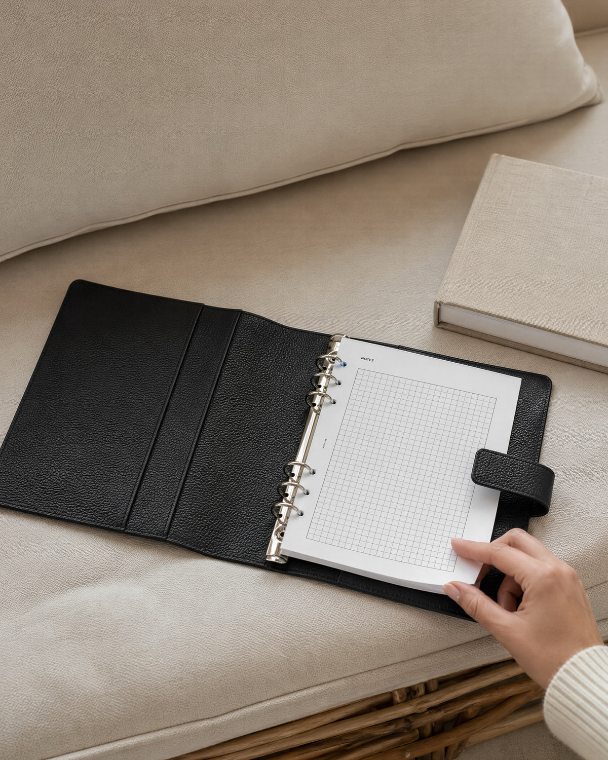 A black textured Filofax leather planner cover with Notairé lined A5 inserts inside, shown open on a beige sofa. A hand is holding the notepad, and a neutral-colored book rests nearby.