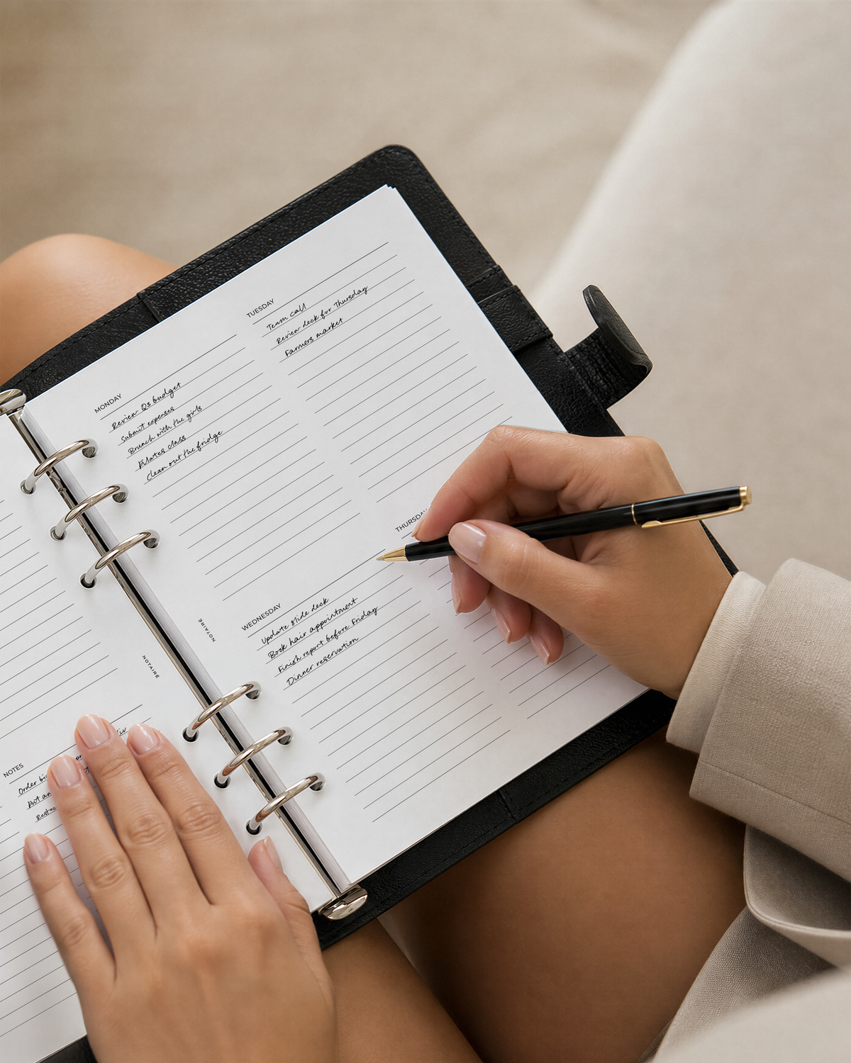 A top-down view of a woman writing in a black Notairé planner with lined pages. The person's hands and arms are visible, resting on their lap, with a beige sofa in the background.