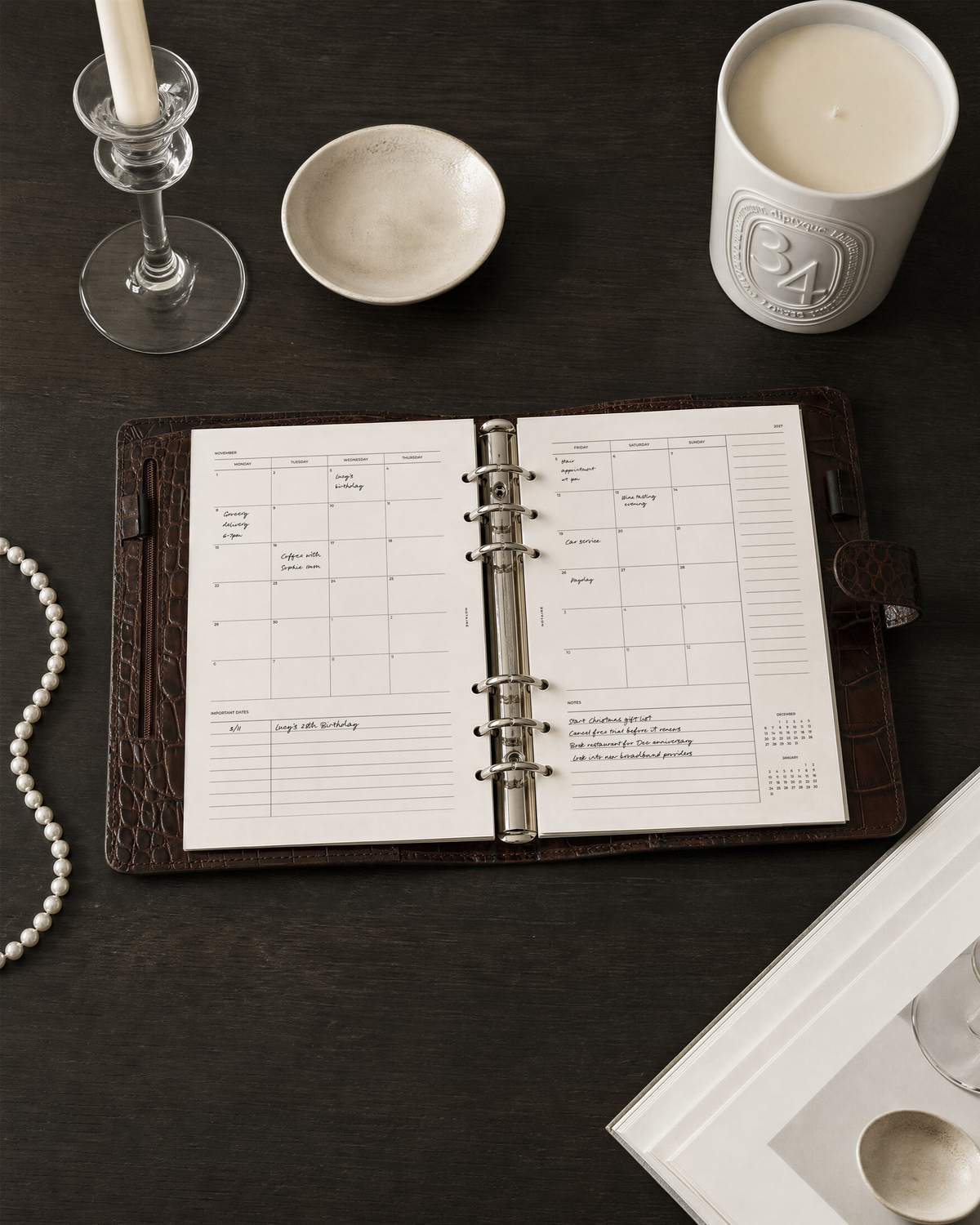 An open ring-bound Filofax Croc agenda  with a brown cover and white Notairé  monthly overview calendar pages, styled on a dark tabletop with candles, a small bowl, a candle jar, and a pearl strand.