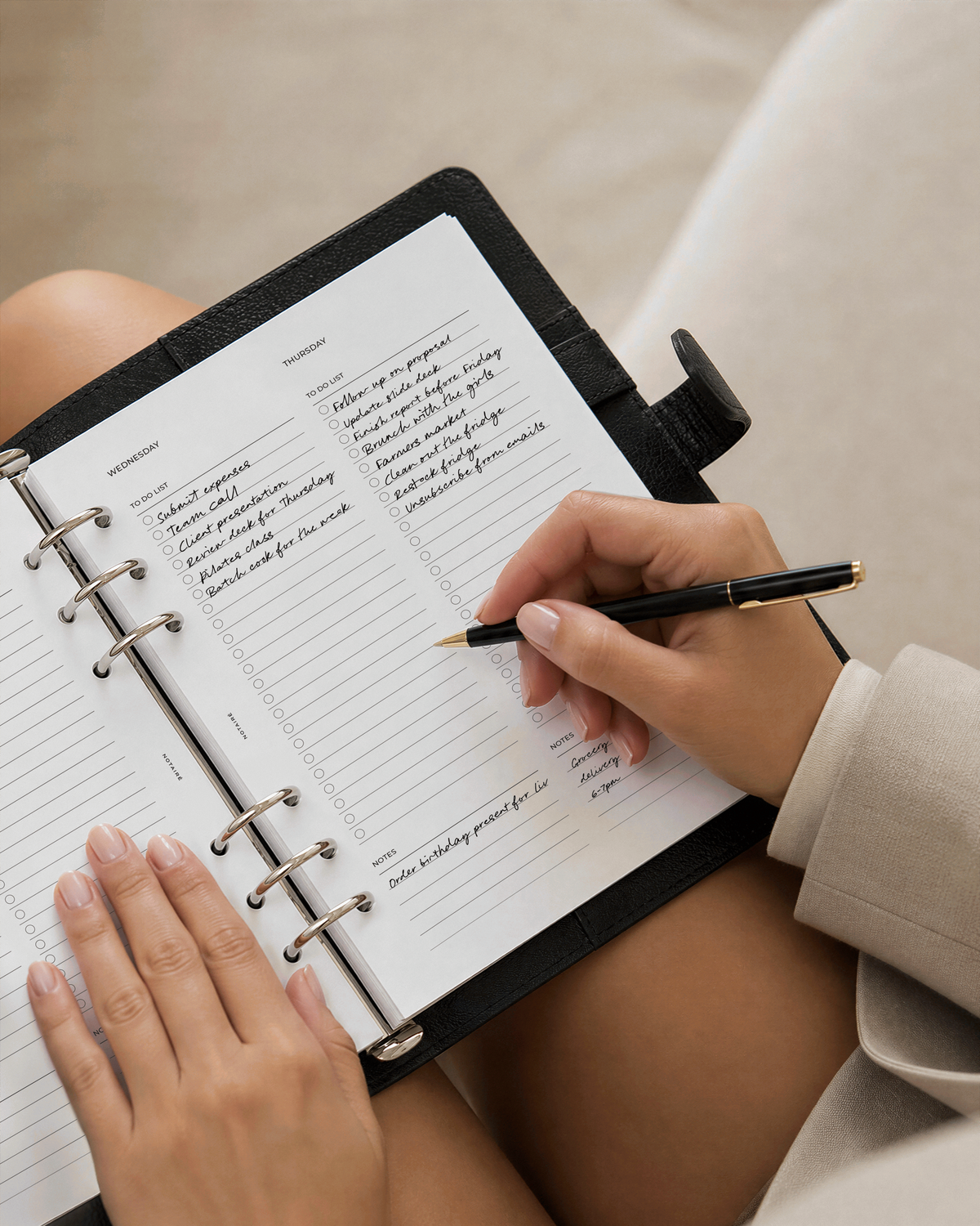 A close-up of a person's hands writing in a black ring-bound planner with lined Notaire Weekly pages, held against a soft beige background.