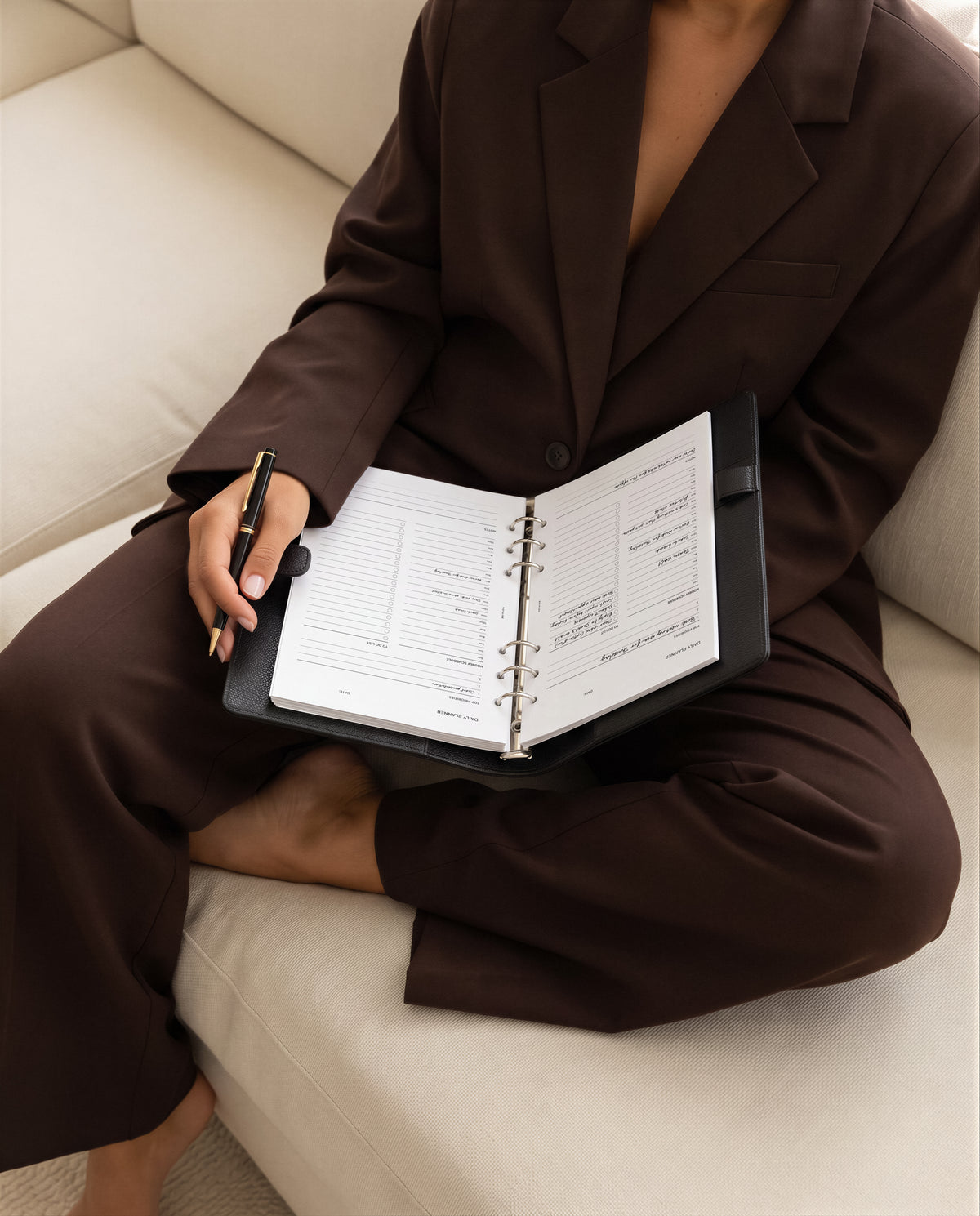 A person in a dark brown tailored suit sits on a cream sofa holding an open black ring binder Filofax planner with a pen with Notairé daily planner inserts inside.