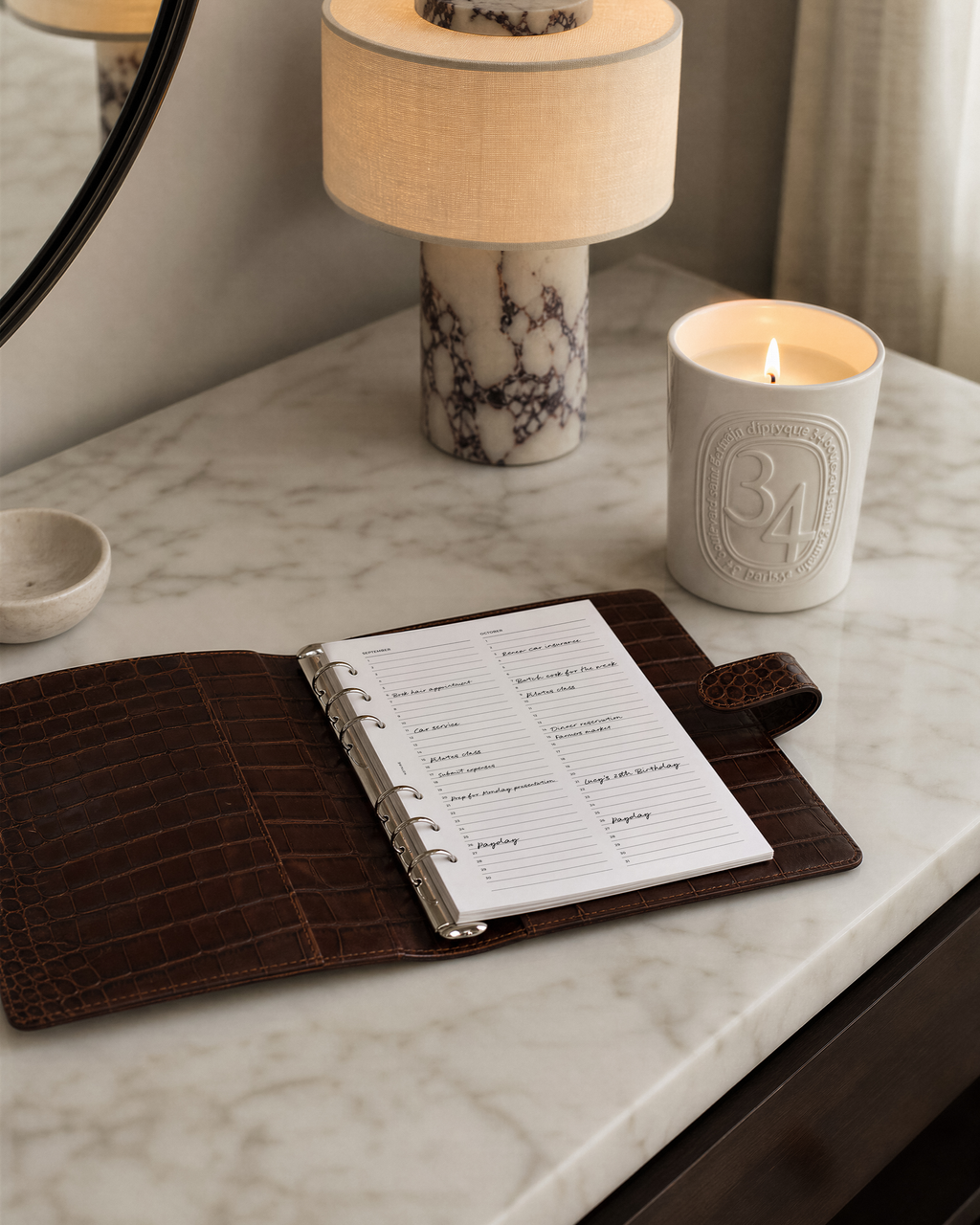 A dark brown Filofax crocodile-embossed leather planner cover open on a white marble desk, holding a white ring-bound agenda with printed Notairé weekly planner inserts. A lit candle, a cream lamp, and part of a mirror are visible in the softly lit room setting.