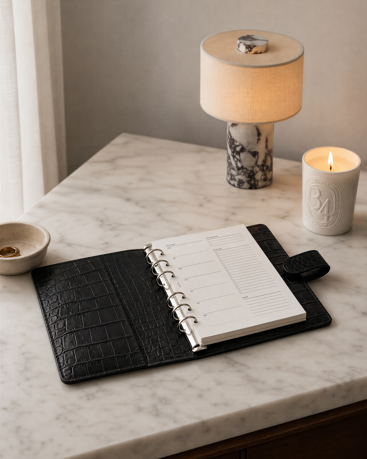A black Filofax crocodile-embossed leather planner cover holding an open Notairé weekly planner with white pages, styled on a white marble tabletop next to a lit Diptyque candle, a small bowl, and a lamp.