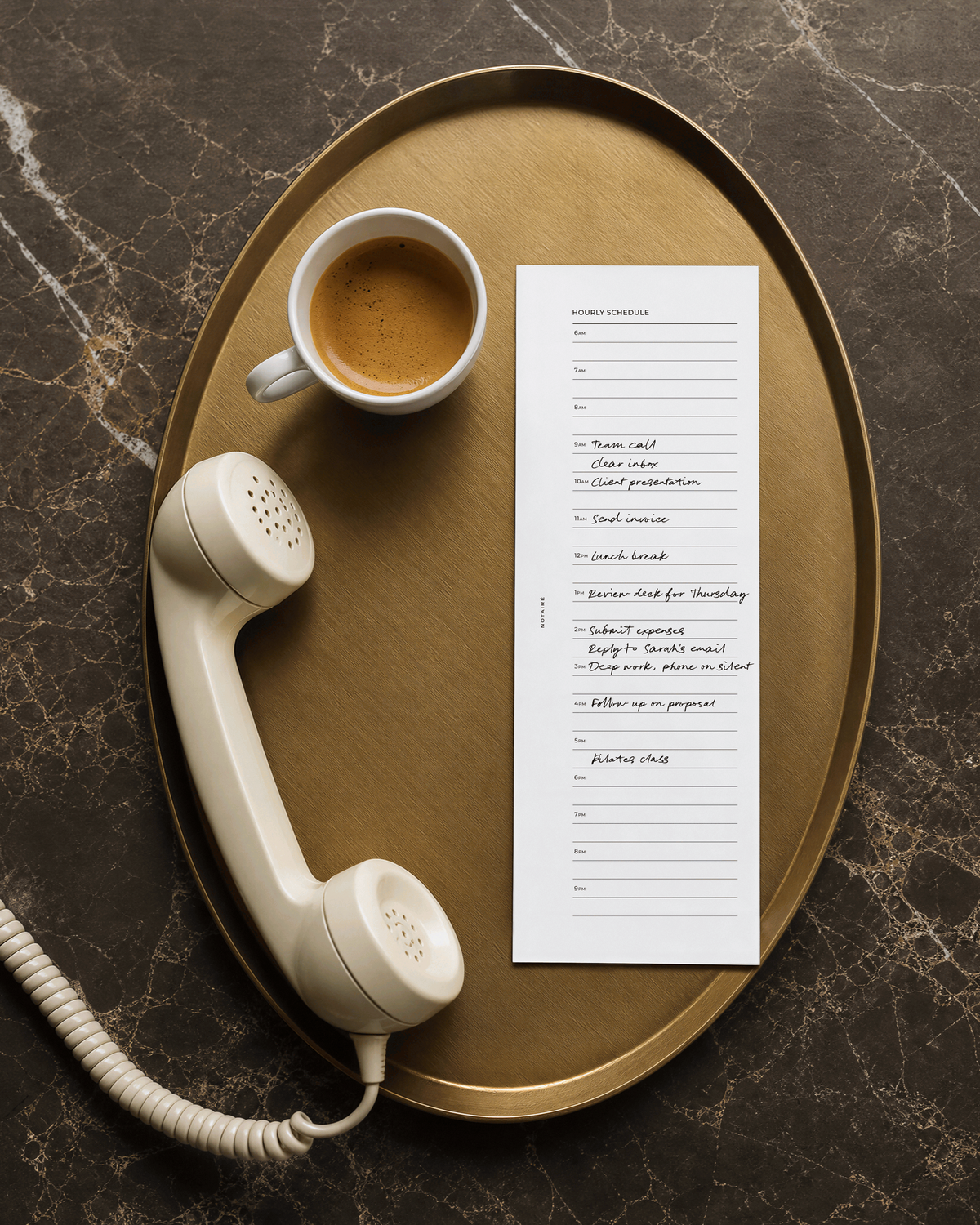 A styled flat-lay featuring a white lined half-page A5 Notairé insert with handwritten text, a vintage cream rotary telephone, and a cup of espresso on an oval gold tray over a dark marble surface.