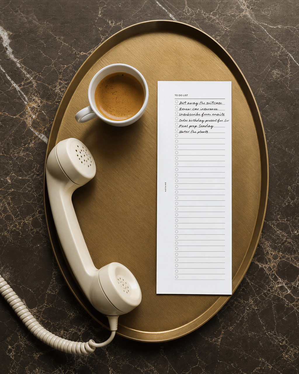 A styled flat-lay featuring a white lined Notairé half-page planner insert with handwritten text, a vintage cream-colored telephone receiver on a coiled cord, and a cup of espresso on an oval gold tray, all set on a dark marble surface.