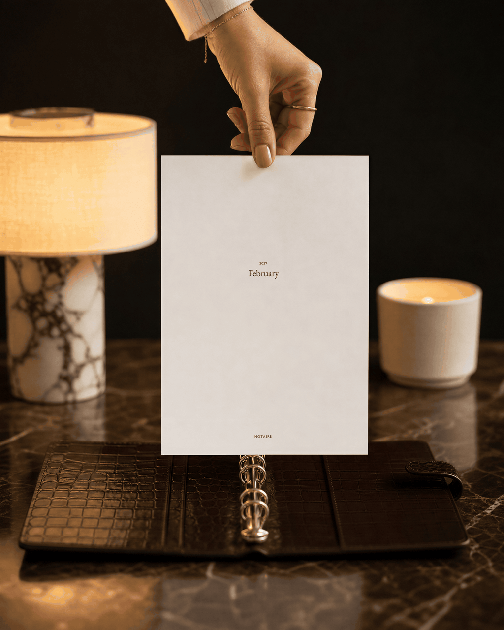 A hand holds a white paper cover for a February ring binder Notairé divider over an open dark brown leather planner on a marble surface, with a lit lamp and candle in the background.