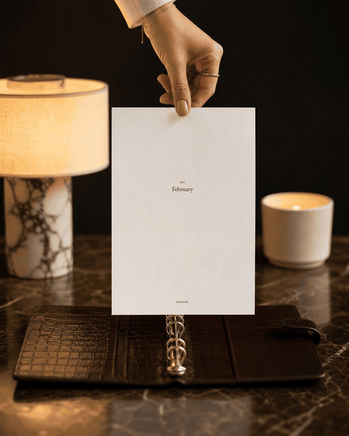 A hand holds a white paper cover for a February ring binder Notairé divider over an open dark brown leather planner on a marble surface, with a lit lamp and candle in the background.