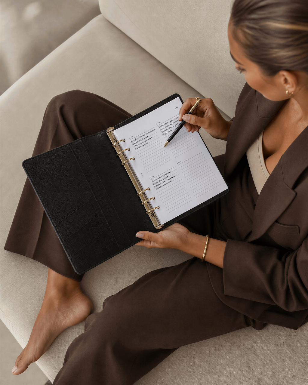 A woman in a brown blazer and pants sits barefoot on a beige sofa, writing in a Filofax planner on Notairé dated planner inserts with a pencil.