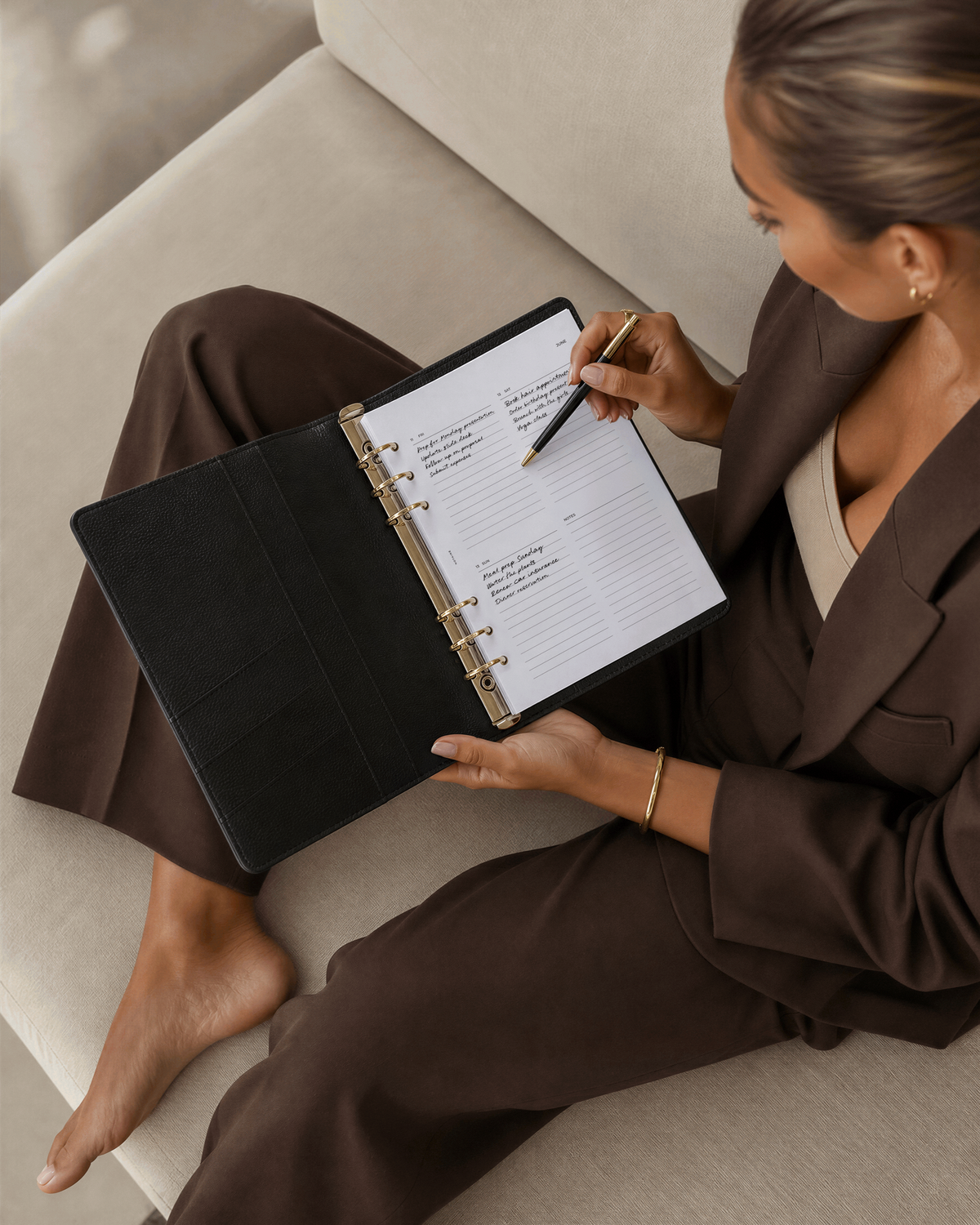 A woman in a brown blazer and pants sits barefoot on a beige sofa, writing in a Filofax planner on Notairé dated planner inserts with a pencil.