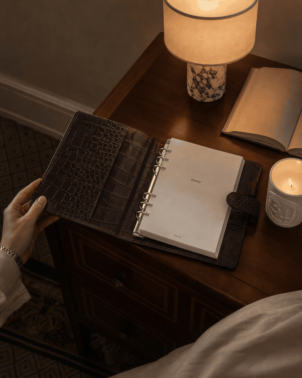 A dark brown crocodile-embossed Filofax  leather planner binder with a white ring-bound Notairé inserts inside, held open on a wooden desk next to a lit lamp, candle, and an open book.