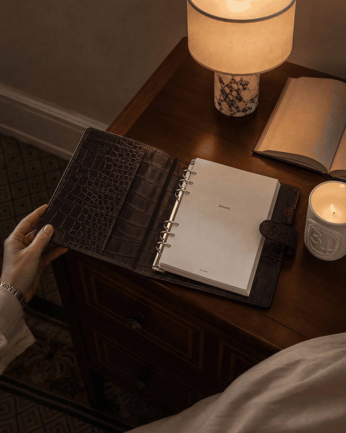 A dark brown crocodile-embossed Filofax  leather planner binder with a white ring-bound Notairé inserts inside, held open on a wooden desk next to a lit lamp, candle, and an open book.
