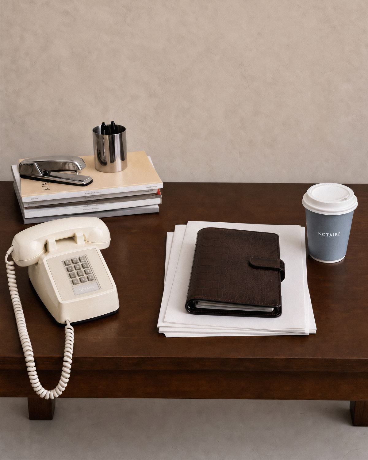 A dark brown textured leather planner or notebook with a black elastic closure, resting on a stack of white papers on a dark wooden table. The scene is styled with a cream rotary telephone, a stack of books with a silver stapler, a metal cup, and a blue coffee cup with the brand 'Notairé' visible on the sleeve.