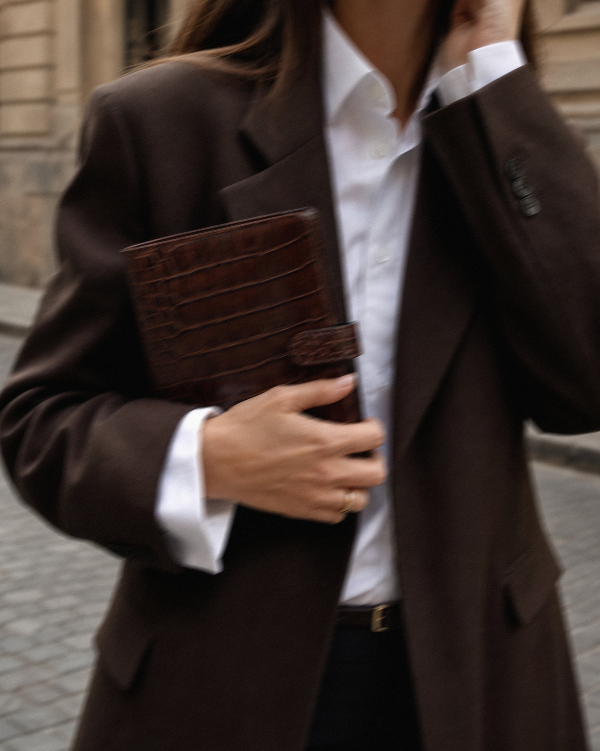 A woman wearing a dark brown blazer over a white collared shirt, holding a dark brown crocodile Filofax agenda with Notairé inserts in an outdoor urban setting.