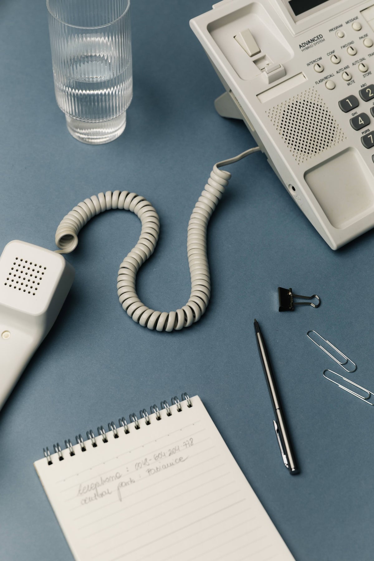 White office phone, pen, notepad, and glass of water on a blue surface