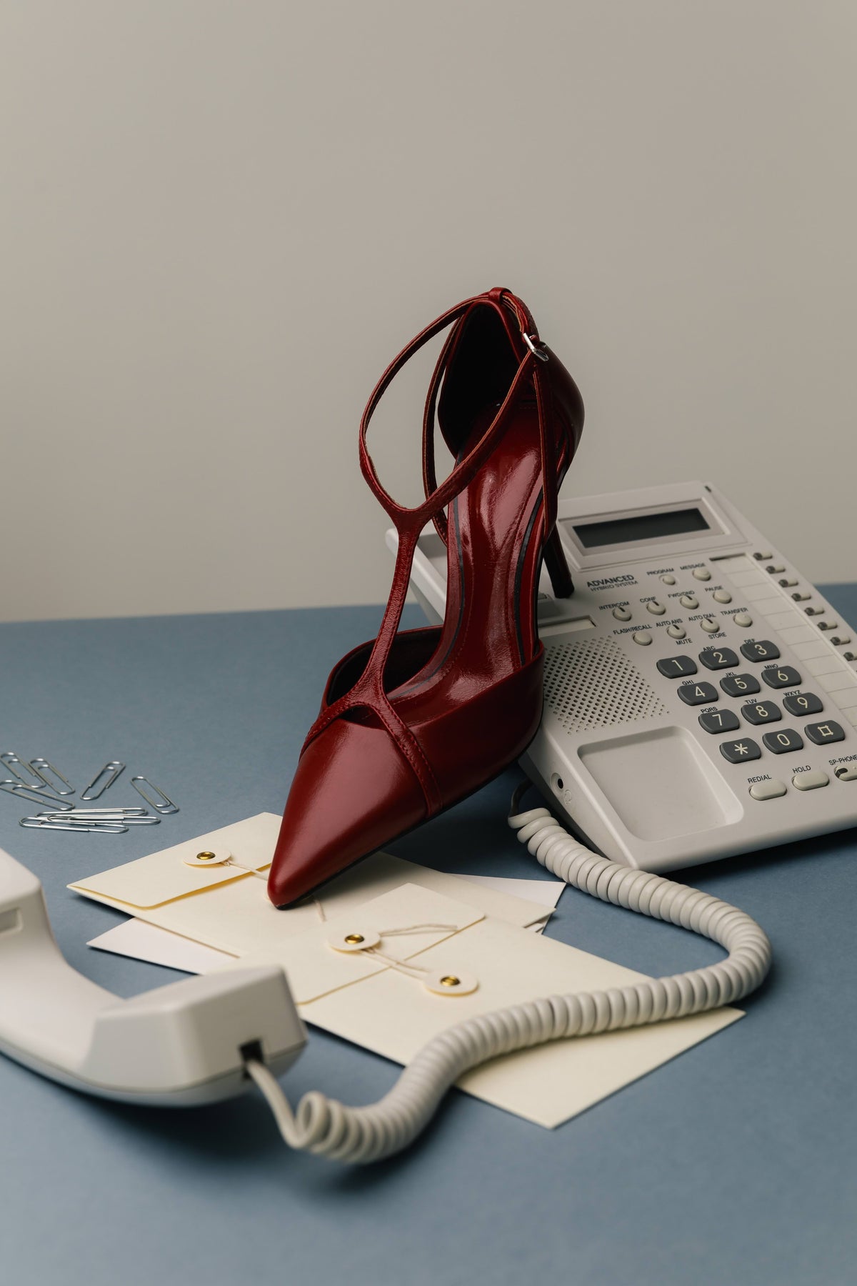 A single pointed-toe high heel in glossy deep burgundy red is styled on a blue tabletop with a white rotary phone, a beige landline, envelopes, and paper clips against a plain light gray wall.